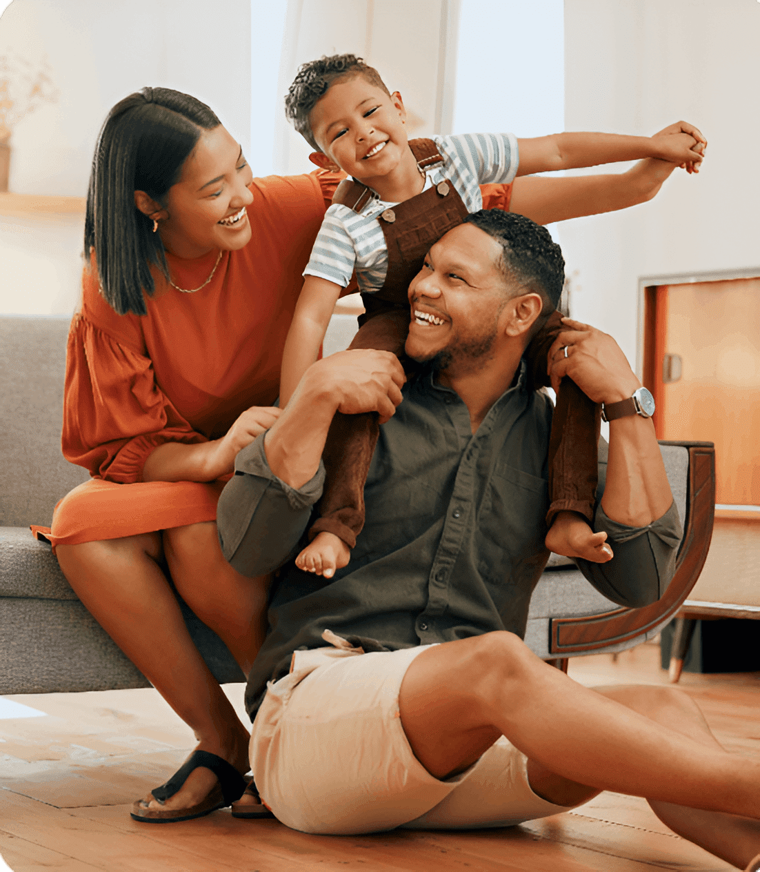 New Project (1) (1) A smiling family of three is indoors. A man is sitting on the floor holding a child on his shoulders, while a woman kneels beside them.