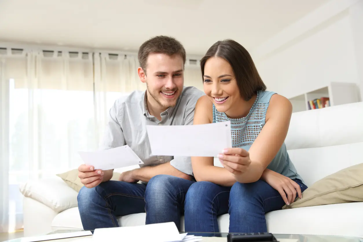A smiling couple sitting on a couch looks at papers together in a bright living room.